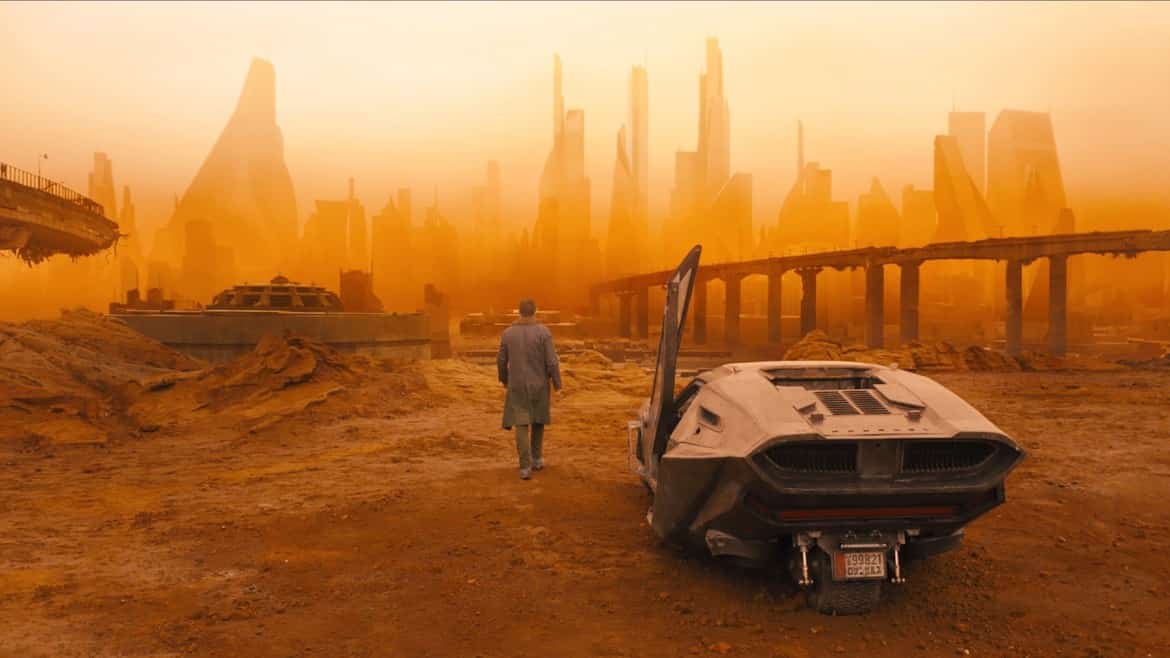 A man with his back towards the camera stands in a desolate landscape with ruins and a giant holographic advertisement in the background